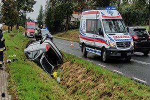 Po lewej stronie drogi w rowie na dachu leży samochód, obok na drodze stoi karetka pogotowia.