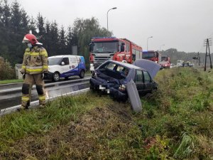 Strażak obok uszkodzonego na poboczu forda. Na drugim planie wóz straży pożarnej.