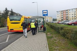 Autobus na przystanku. Na chodniku pasażerowie i policjant.