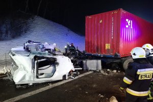 Kabina pojazdu lezy na jezdni. Za nią stoi naczepa a obok strażacy