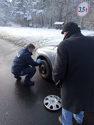 Policjant wymienia koło w samochodzie. Obok pojazdu tyłem stoi mężczyzna.