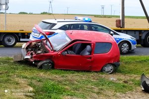 Uszkodzony czerwony samochód stoi na poboczu. Za ni widać stojący radiowóz.