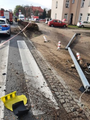 Przejście dla pieszych. Na chodniku leży wyrwany sygnalizator. N a drugim planie widać pojazdy służb ratunkowych oraz uszkodzony czerwony samochód