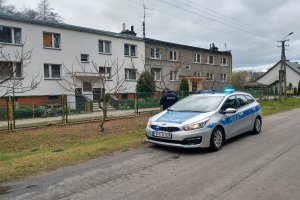 Policjant stoi obok radiowozu. Na drugim planie znajduje się budynek mieszkalny.