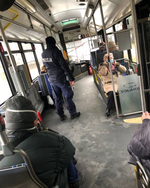 Policjant stojący w autobusie. Po bokach siedzą pasażerowie