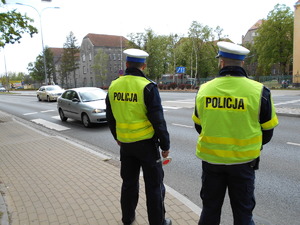 dwóch policjantów stoi w pobliżu przejścia dla pieszych. W tle widać jadący samochód osobowy