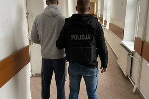 policjant w czarnej kamizelce z napisem policja prowadzi zatrzymanego, obaj odwróceni tyłem