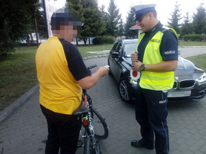 na zdjęciu z lewej strony stoi mężczyzna w pomarańczowo-czarnej koszulce, przed nim stoi rower. Z prawej strony stoi policjant w odblaskowej kamizelce i jest zwrócony w stronę rowerzysty