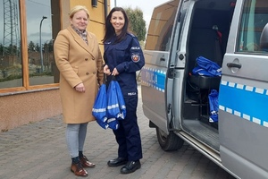 na zdjęciu z lewej strony stoi kobieta w beżowym płaszczu. Obok tej kobiety stoi policjantka i trzyma w rękach niebieskie worki z upominkami. Z prawej strony widać radiowóz policyjny.