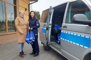 przed budynkiem stoi kobieta w beżowym płaszczu, obok niej stoi policjantka i trzyma w rękach niebieskie worki z upominkami. Z prawej strony widać radiowóz policyjny