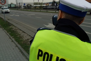 policjant trzyma w ręku urządzenie do pomiaru prędkości. W tle widać jadący pojazd