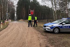 przy przejeździe kolejowym stoi policjant i funkcjonariusz straży ochrony kolei, przy tym przejeździe umieszczony jest znak drogowy STOP. Na zdjęciu z prawej strony widać radiowóz policyjny