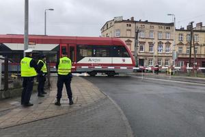 przed przejazdem kolejowym stoi policjant i dwóch funkcjonariuszy ochrony kolei, mają założone kamizelki odblaskowe. Zapory na przejeździe są opuszczone. Widać jadący szynobus.