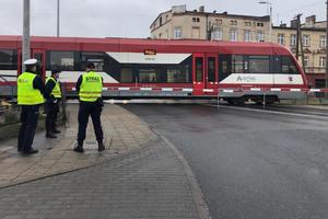 przed przejazdem kolejowym stoi policjant i dwóch funkcjonariuszy straży ochrony kolei, mają założone kamizelki odblaskowe. Zapory na przejeździe są opuszczone, Widać jadący szynobus.