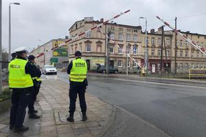 przed przejazdem kolejowym z lewej strony stoi policjant i dwóch funkcjonariuszy straży ochrony kolei, mają założone kamizelki odblaskowe. Widać jak zapory są opuszczane. Po drugiej stronie przejazdu kolejowego stoi samochód osobowy koloru siwego. W tle widać kamienice.