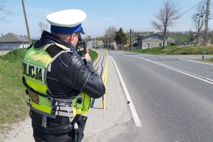 policjant z ruchu drogowego stoi tyłem, ma założoną kamizelką odblaskową z napisem policja, na głowie ma czapkę w kolorze białym. Policjant stoi na poboczu drogi i mierzy prędkość urządzeniem pomiarowym. W oddali widać jadący pojazd.