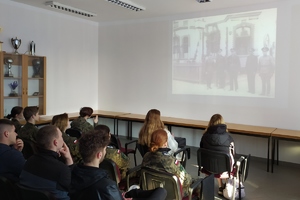 na zdjęciu widać grupę osób siedzących na krzesłach w auli. Osoby są odwrócone tyłem i patrzą na ścianę na której wyświetlane są historyczne czarno białe zdjęcia, które przedstawiają policjantów w mundurach.