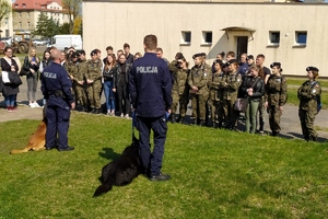 na zdjęciu widać dwóch policjantów ubranych w mundur koloru granatowego z tyłu na plecach widać napis Policja. Z lewej strony tych policjantów siedzą na trawie psy służbowe. Przed policjantami stoi grupa młodych dziewczyn i chłopców, część z nich ubrana jest w mundury wojskowe a część ubrana w odzież cywilną. Młodzież zwrócona jest w stronę policjantów.