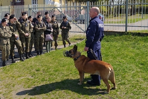 z lewej strony stoi grupa młodych dziewczyn i chłopców, którzy ubrani są w mundur wojskowy. Z prawej strony przed tymi młodymi osobami stoi policjant w granatowym mundurze, który trzyma na smyczy z lewej strony psa służbowego. Policjant zwrócony jest to młodzieży.