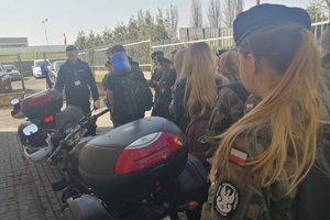 z lewej strony przy ścianie budynku stoją dwa motocykle policyjne odwrócone tyłem. Za motocyklami stoi policjant w mundurze koloru granatowego. W stronę policjanta zwrócone są młode dziewczyny i chłopcy. Część z nich ubrana jest w mundur wojskowy.