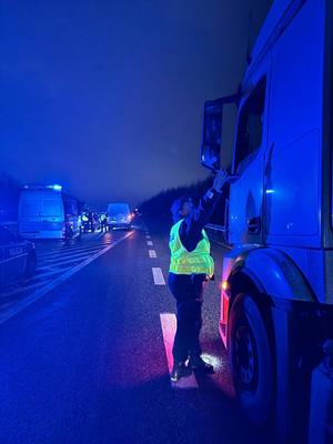 Funkcjonariusz przeprowadza badanie trzeźwości kierującego. Na planie widoczny pojazd ciężarowy oraz działania policjantów w błyskach koloru niebieskiego.