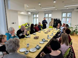 Spotkanie przy stole: Grupa osób siedzi przy długim stole nakrytym żółtym obrusem, na którym stoją pączki, ciasto i kawa. Na środku stoi policjant w mundurze i przemawia do zebranych.