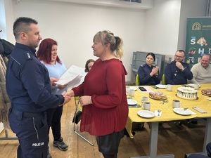 Uścisk dłoni: Policjant w ciemnym mundurze podaje rękę kobiecie w bordowej bluzce. W tle widać pozostałych uczestników spotkania siedzących przy stole z poczęstunkiem.