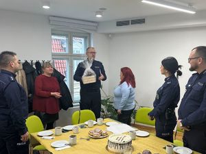 Wręczanie prezentu: Policjant stoi na środku i trzyma zapakowany w folię kosz upominkowy, zwracając się do kobiety w bordowej bluzce. Na stole wciąż widoczne są pączki i tort.