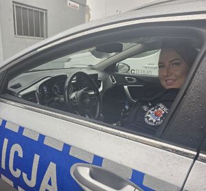 Uśmiechnięta kobieta siedzi za kierownicą srebrno-niebieskiego radiowozu z wyraźnym napisem "POLICJA" na boku. Ma na sobie ciemny mundur z naszywką polskiej policji na ramieniu. Auto jest mokre od deszczu, a w tle widać bloki mieszkalne.