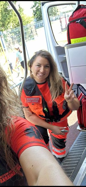 Kobieta w pomarańczowym mundurze ratownika medycznego (z napisem "RATOWNIK" na piersi) uśmiecha się i pokazuje palcami znak "V" (zwycięstwo/pokój). Zdjęcie jest zrobione z perspektywy wnętrza karetki, w której widoczne są czerwone torby medyczne i sprzęt. W tle, przez otwarte drzwi pojazdu, widać rozmyte sylwetki dzieci na zewnątrz.