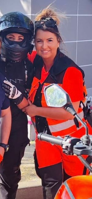 Ta sama kobieta pozuje obok osoby w czarnym kasku motocyklowym. Ma na sobie jaskrawopomarańczowy strój służb ratunkowych (kamizelkę odblaskową i spodnie). Na głowie ma okulary przeciwsłoneczne, a jej włosy są upięte w luźny kok. Na pierwszym planie widoczna jest kierownica i lusterko motocykla.